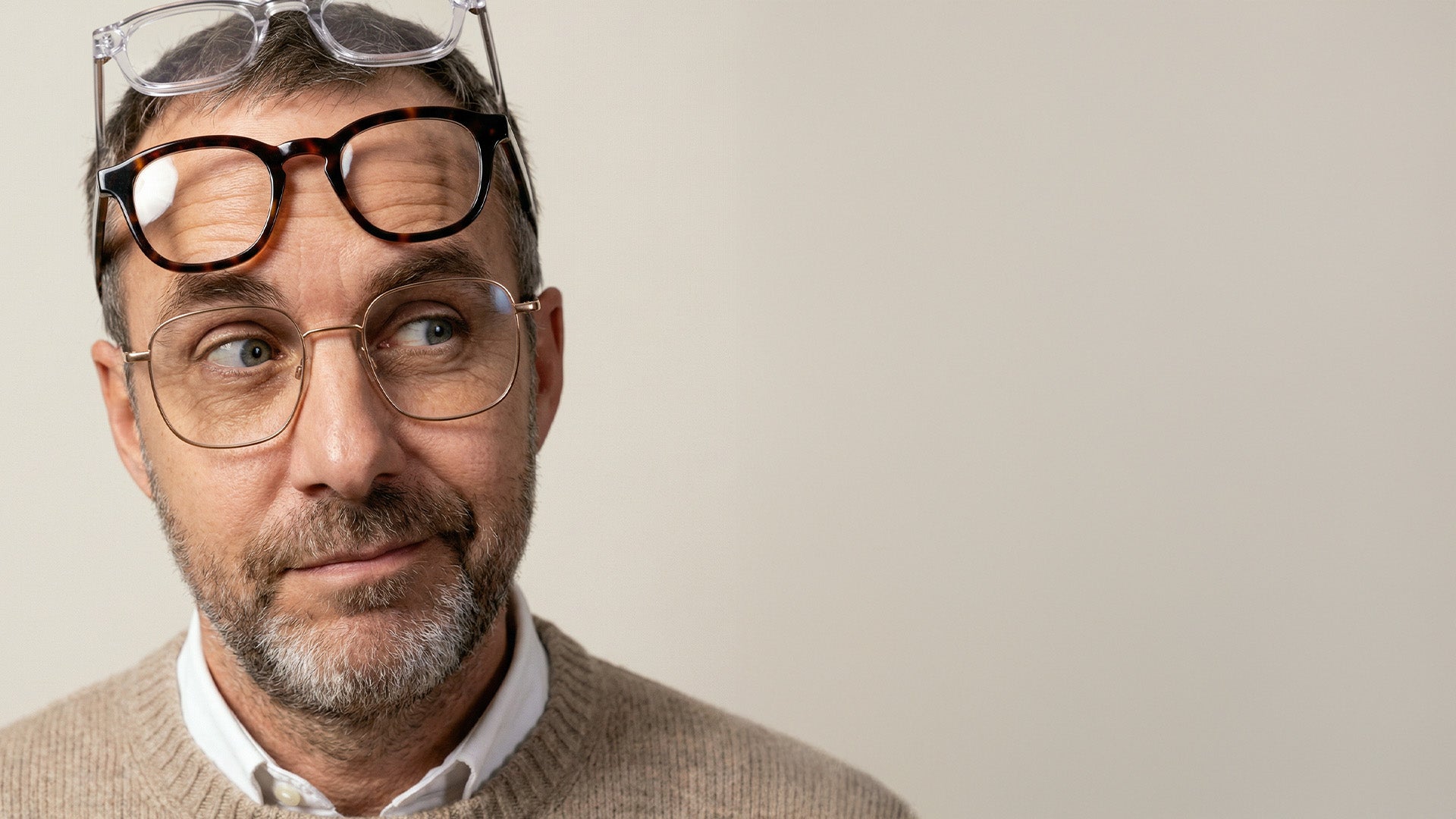 Man wearing glasses with additional pairs on his head, looking to the side. Neutral background, casual attire, thoughtful expression.