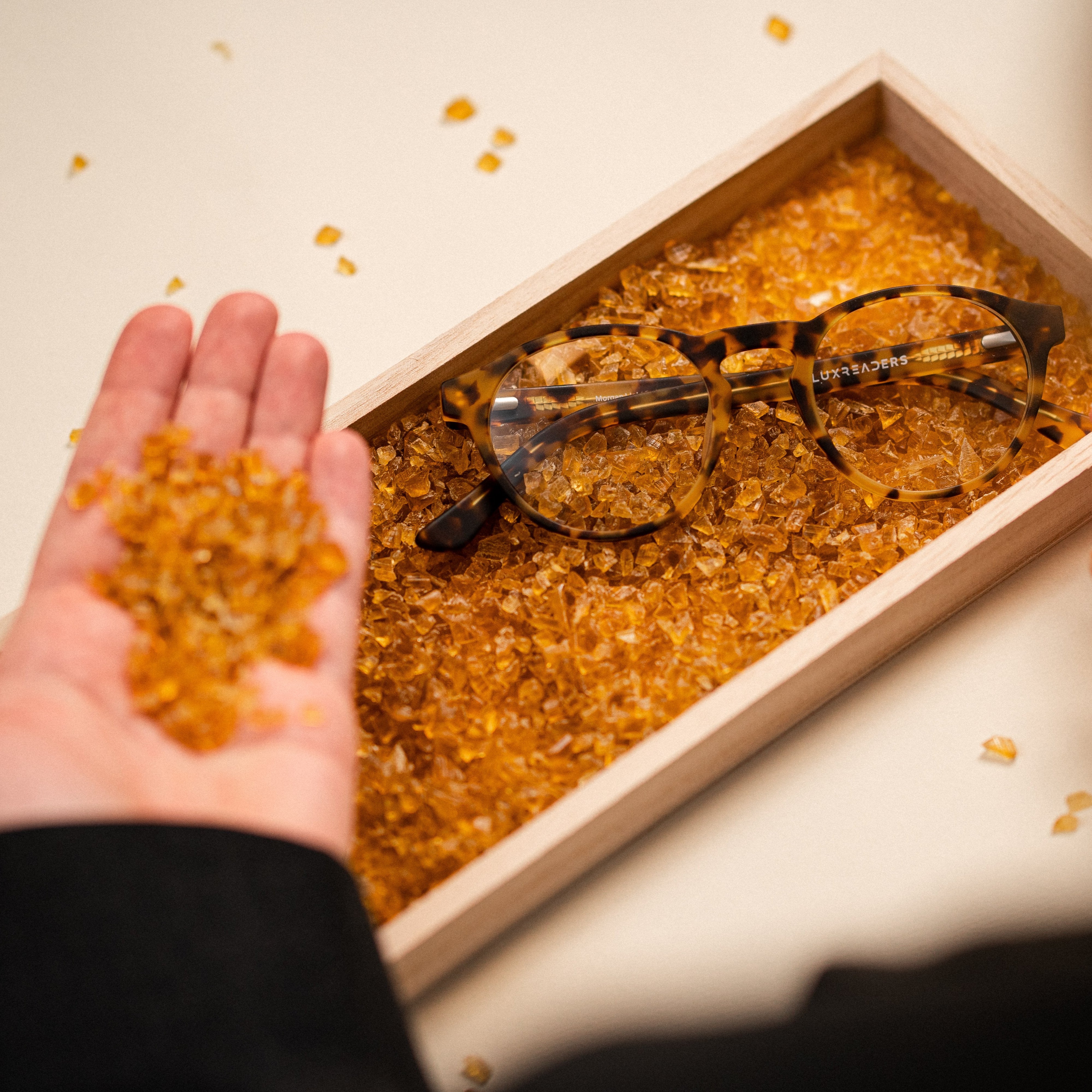Tortoiseshell glasses labeled Luxreaders on amber crystals, one hand holding crystals, in a wooden box.