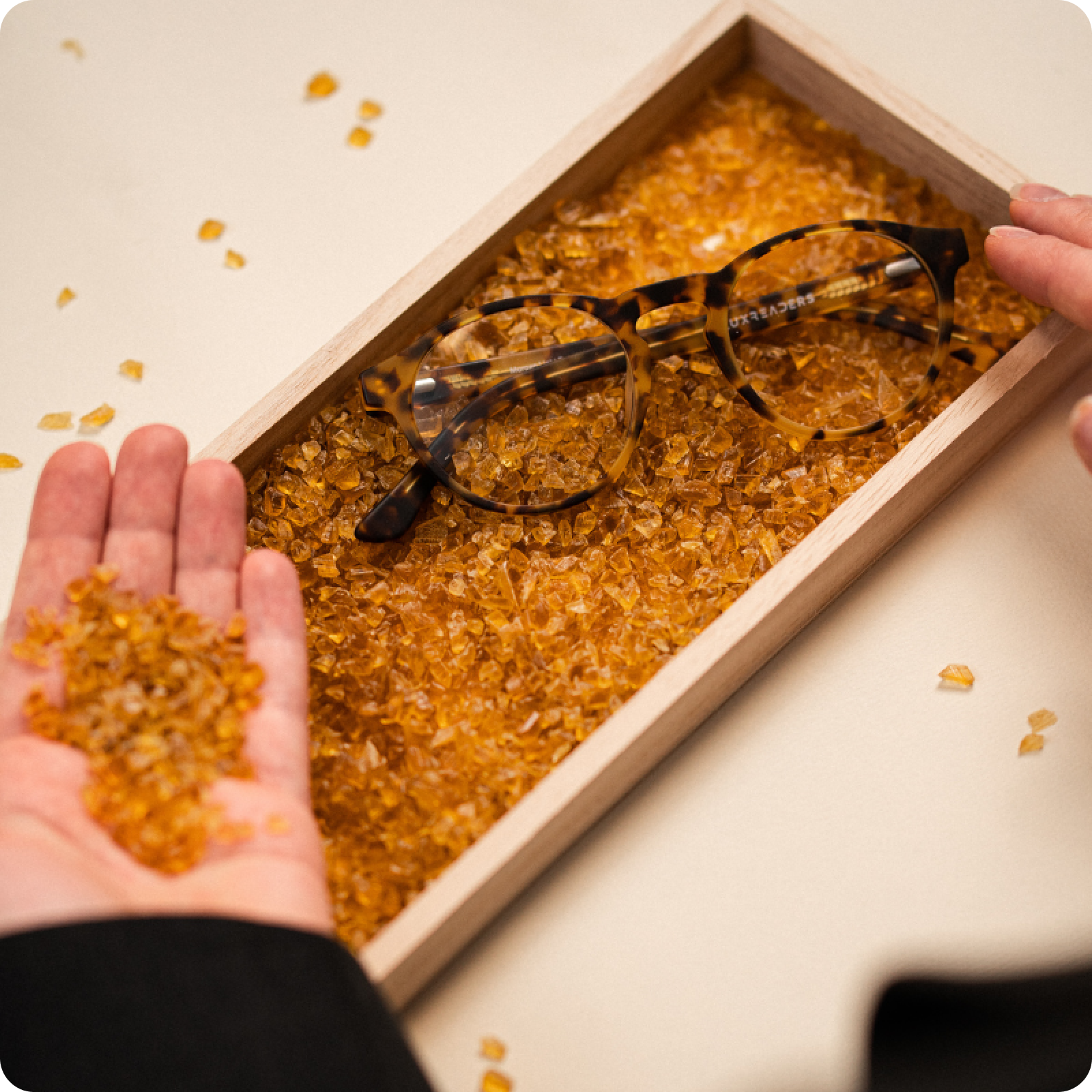 Tortoiseshell glasses on amber crystals in a wooden tray, with a hand holding some crystals.