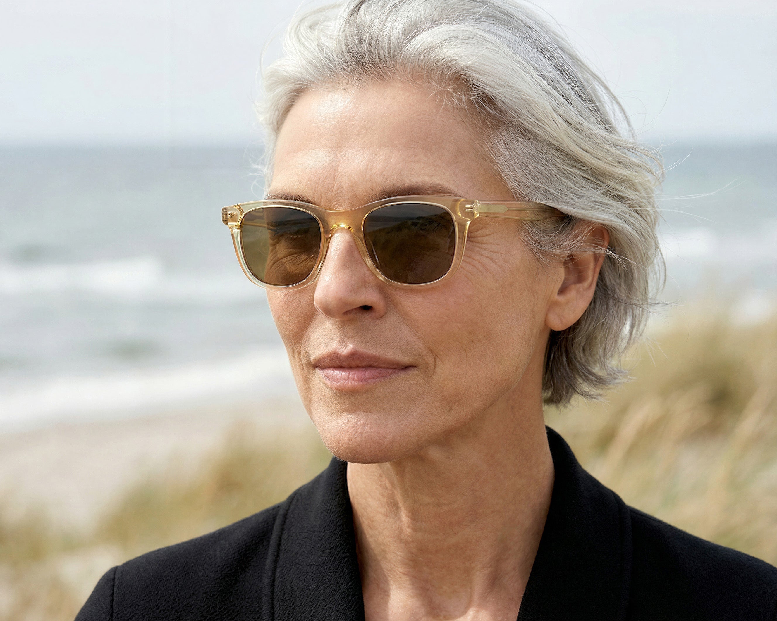 Woman with gray hair wearing sunglasses on a windy beach, gazing into the distance, with ocean waves behind her.