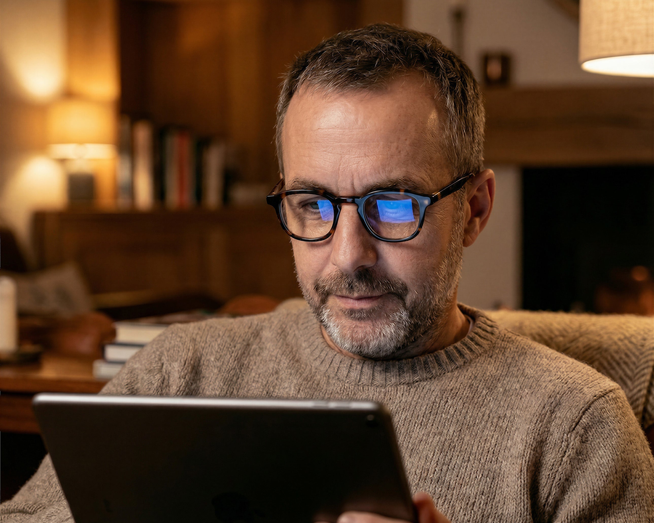 Man wearing glasses, reading a tablet in a cozy, warmly-lit room with bookshelves in the background.