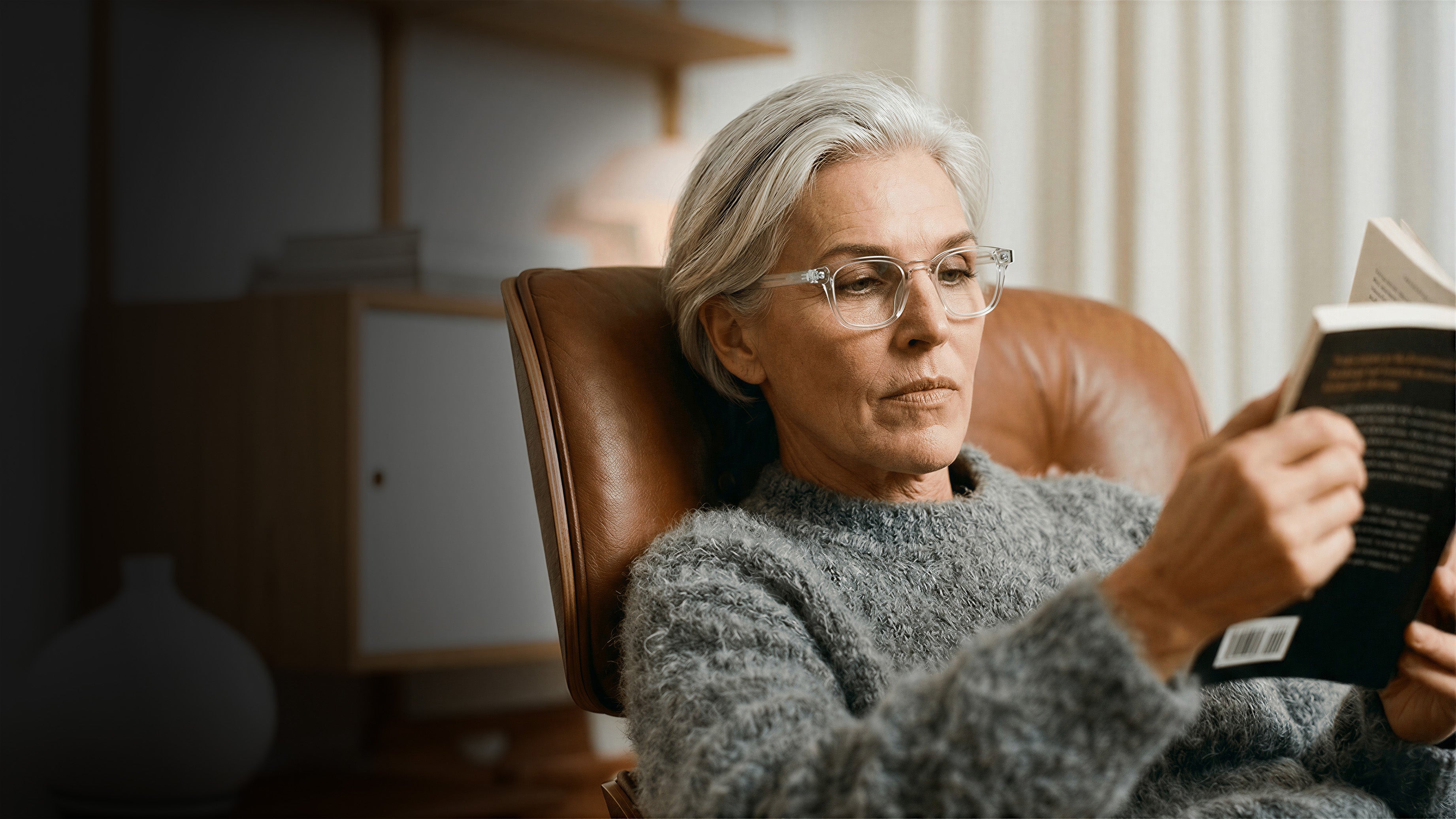 Woman with gray hair wearing eyeglasses and a gray sweater, sitting in a leather chair, reading a book attentively.