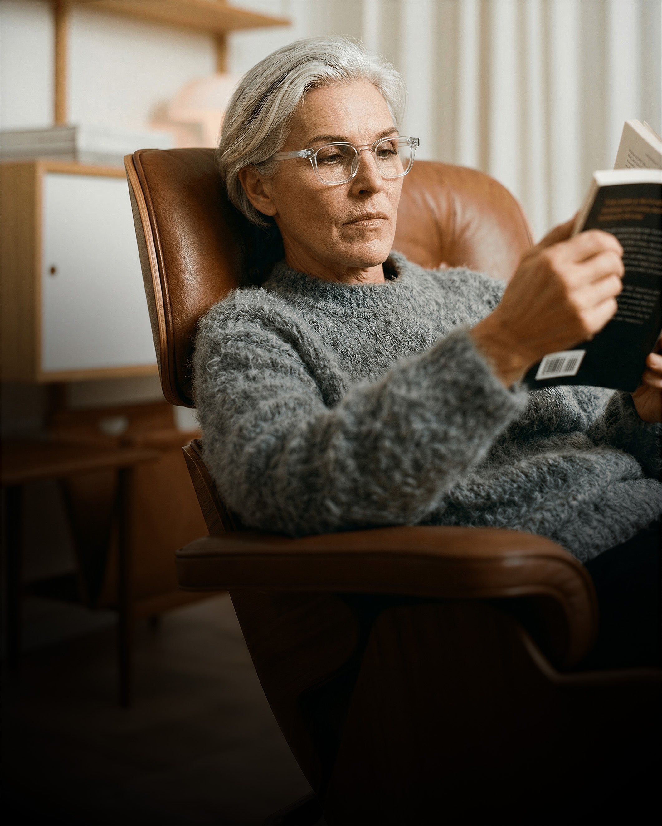 Elderly person with glasses reading a book in a cozy chair, wearing a gray sweater. Warm, soft lighting in a home setting.
