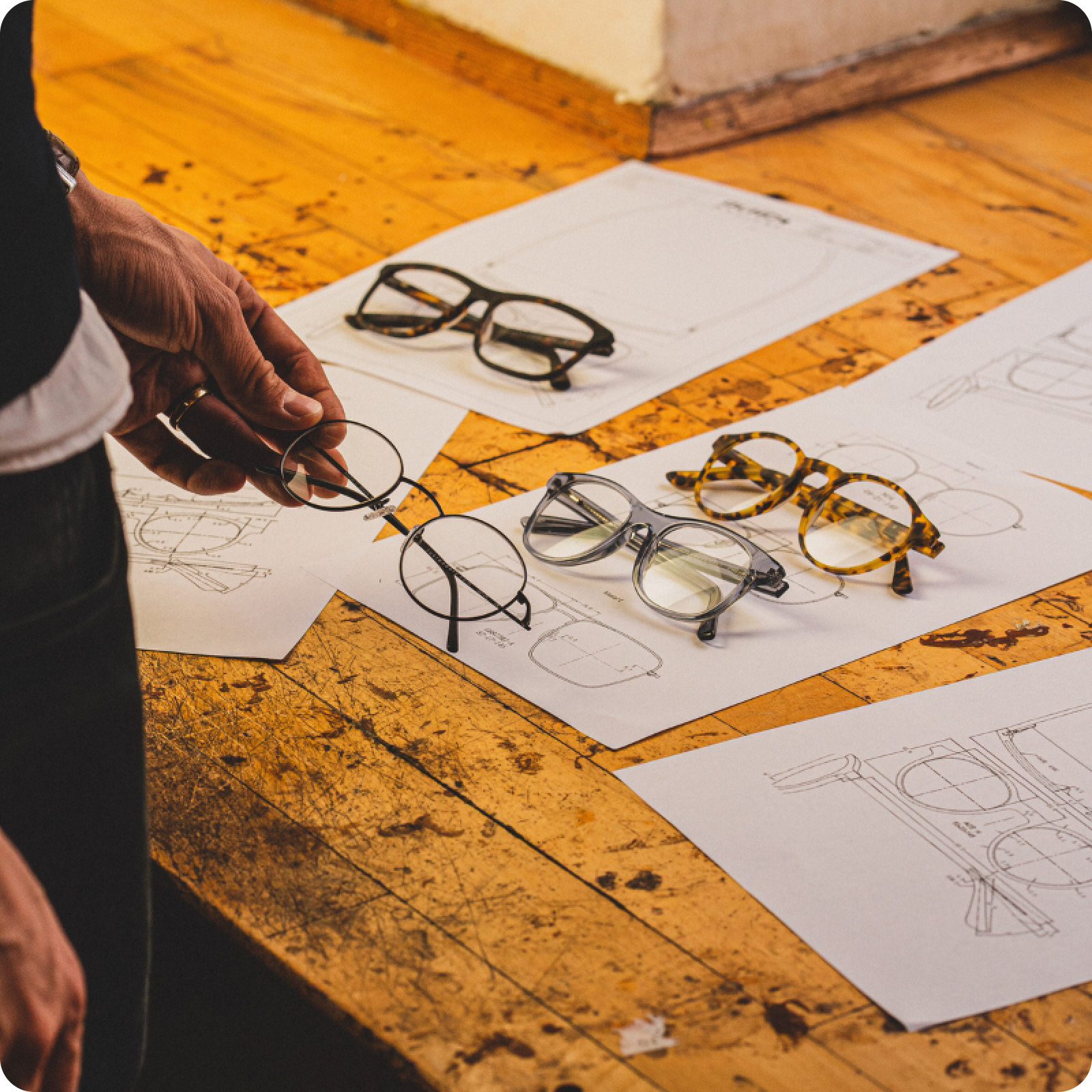 Person holding glasses over a wooden table with four other frames and design sketches, showcasing eyewear selection.