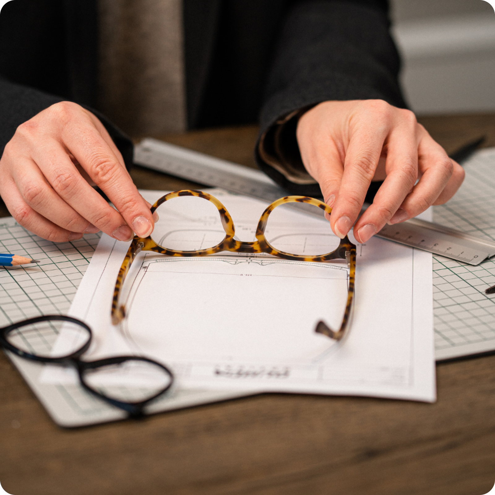 Hands holding tortoiseshell glasses over design sketches, rulers, and a pencil on a wooden desk.